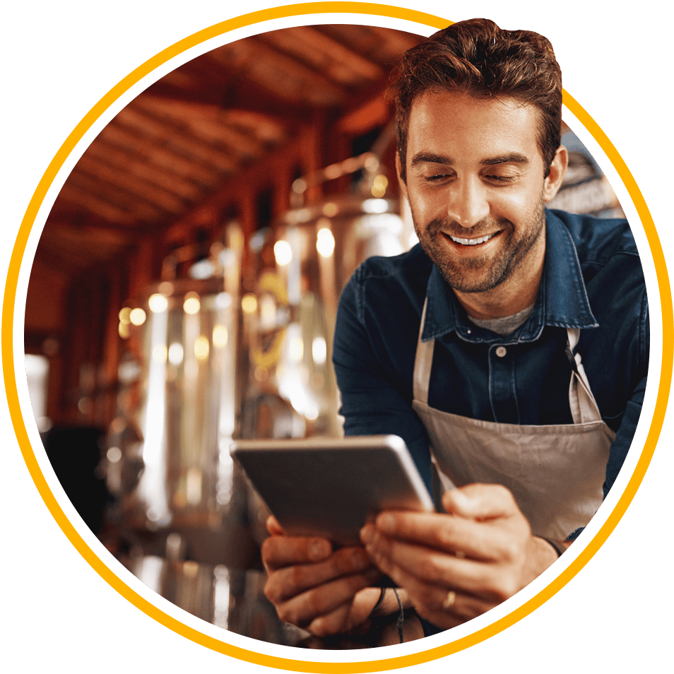 A smiling man wearing an apron looks at data on a tablet in a distillery.