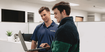 A smiling man shows his male coworker something on his laptop in an office