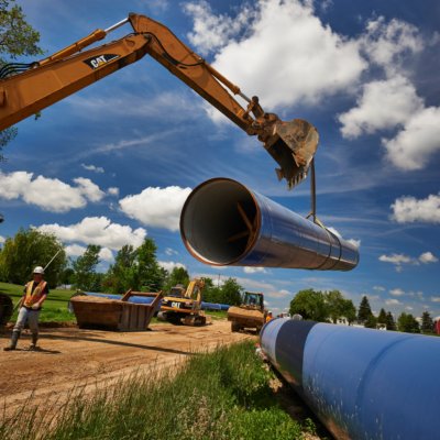 Construction site with one large metal pipe being lifted by heavy machinery, while another metal pipe sits on the grass.