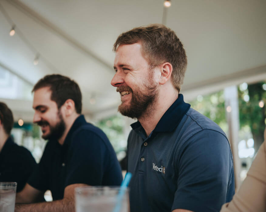 Two smiling Vertech team members, both wearing Vertech polo shirts, sit at a table in an outdoor setting.