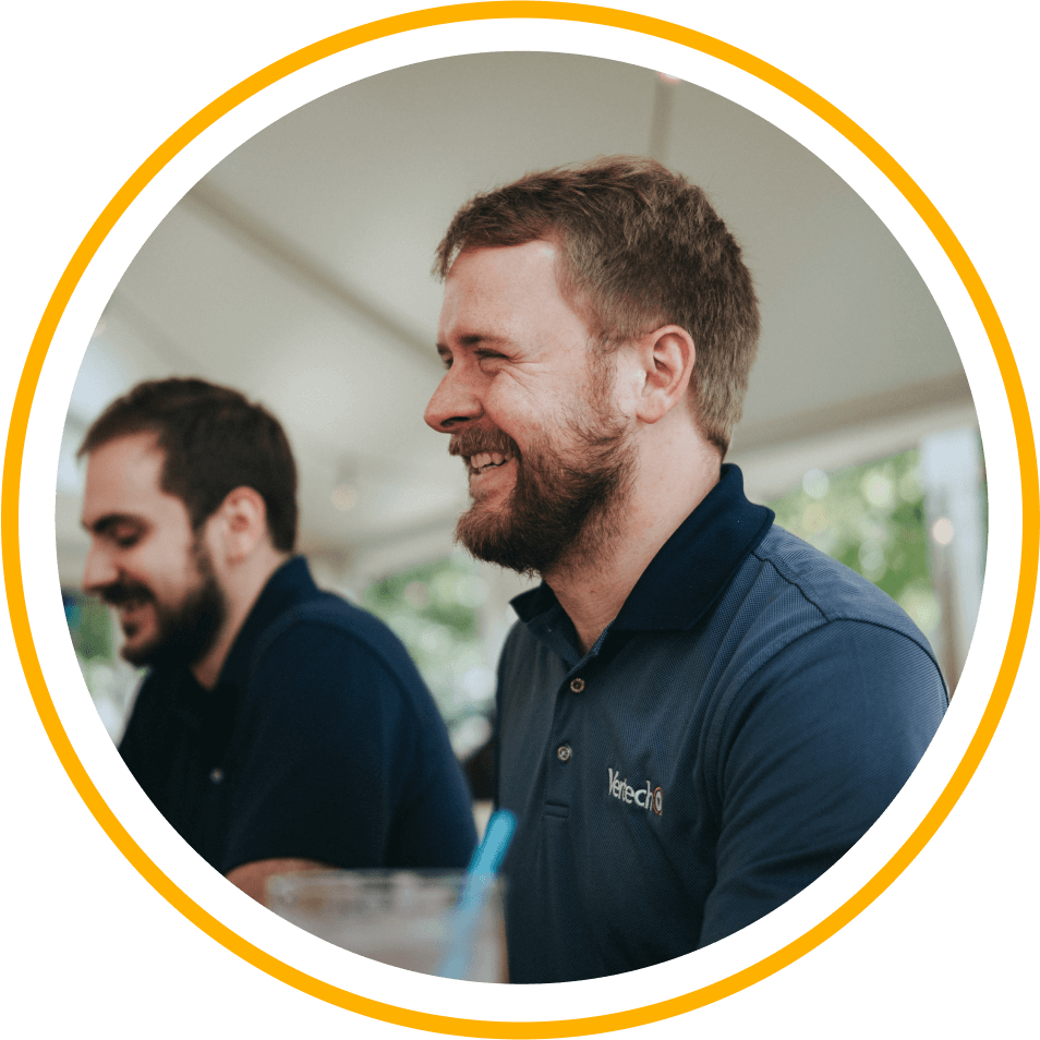 Two smiling Vertech team members, both wearing Vertech polo shirts, sit at a table in an outdoor setting.