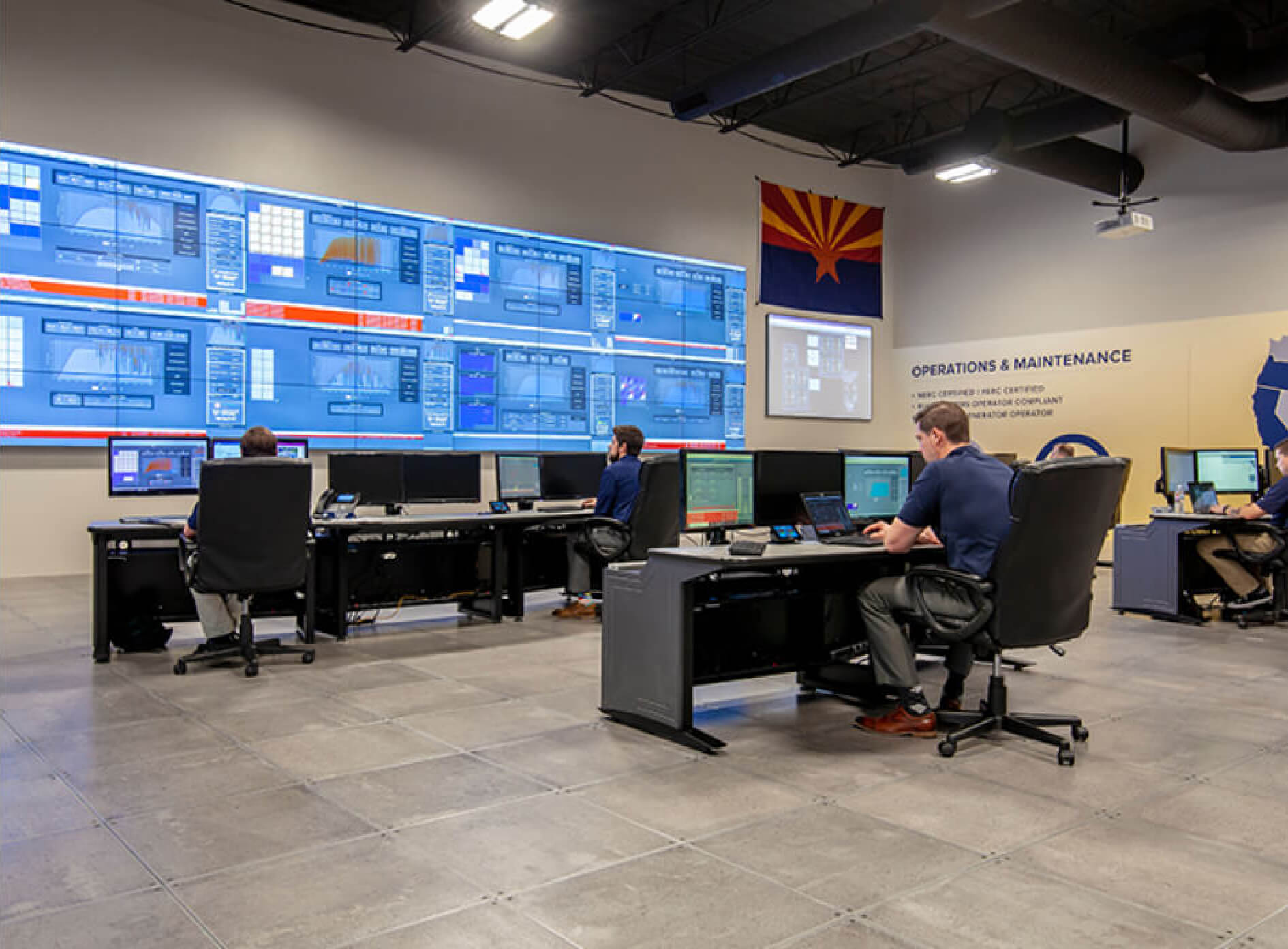 DEPCOM Power operations center, with multiple team members sitting at desks in front of computers and a large projection of various control screens on the wall.