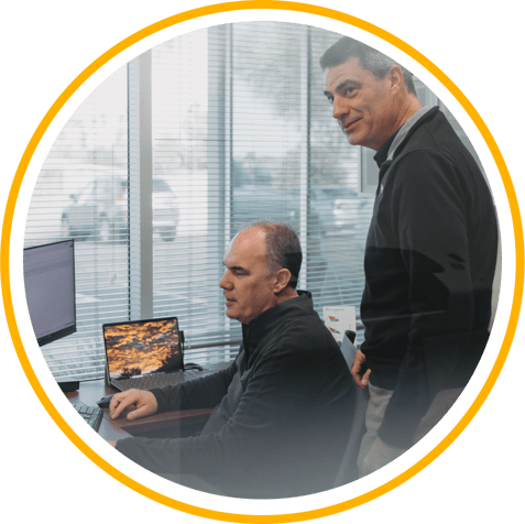 Two Vertech team members in an office, one sitting at a desk in front of a computer, and the other standing behind.