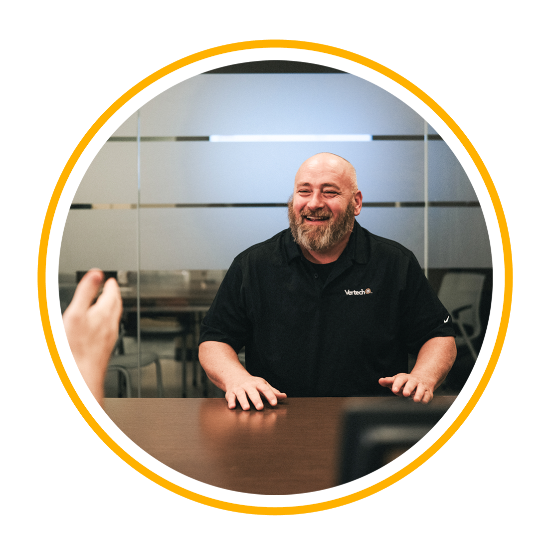 A Vertech team member with a beard wearing a black polo, smiling at someone sitting across from him.