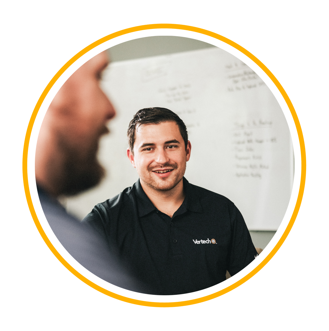 A Vertech team member in a black polo shirt stands in front of a whiteboard, smiling.