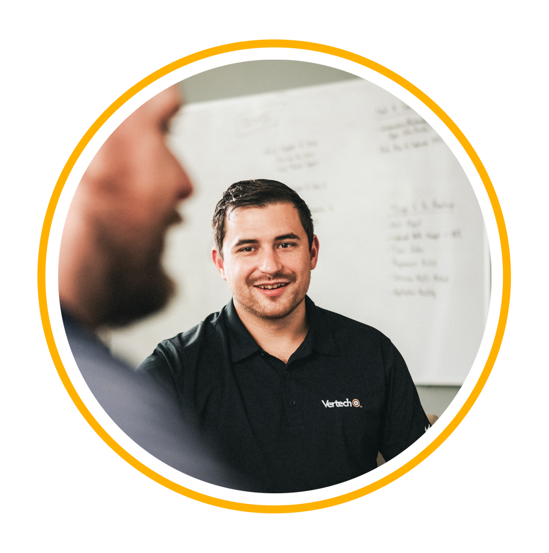 A Vertech team member in a black polo shirt stands in front of a whiteboard, smiling.